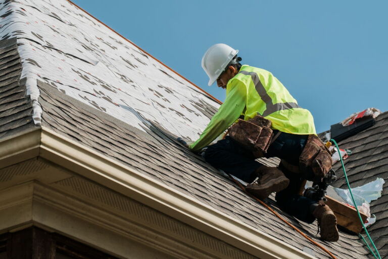 Roofer in a safety vest repairing shingles on a roof under a clear blue sky